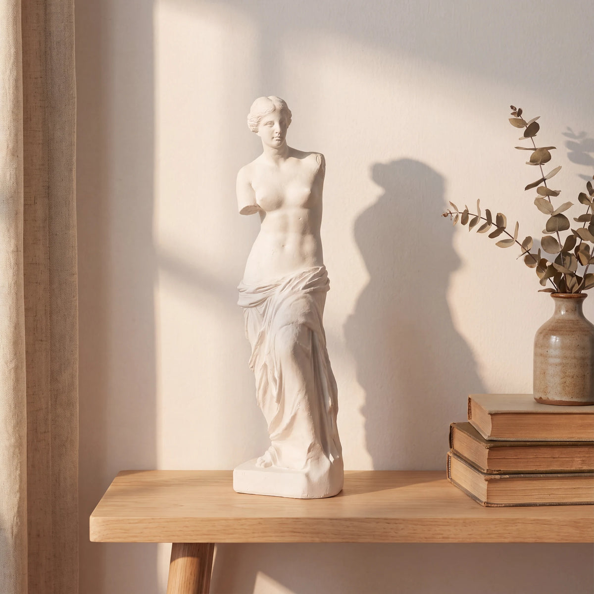 Escultura Vênus de Milo em gesso cru sobre estante de madeira clara com livros e vaso de eucalipto seco