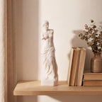 Escultura Vênus de Milo em gesso cru em prateleira de madeira com luz natural suave e cortina de linho
