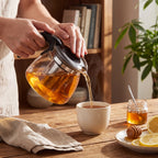 Bule de vidro com infusor servindo chá quente em xícara com mel e limão ao lado