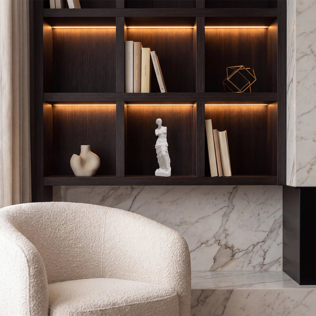 Escultura Vênus de Milo em gesso cru em estante escura de escritório com LED âmbar e poltrona bouclé