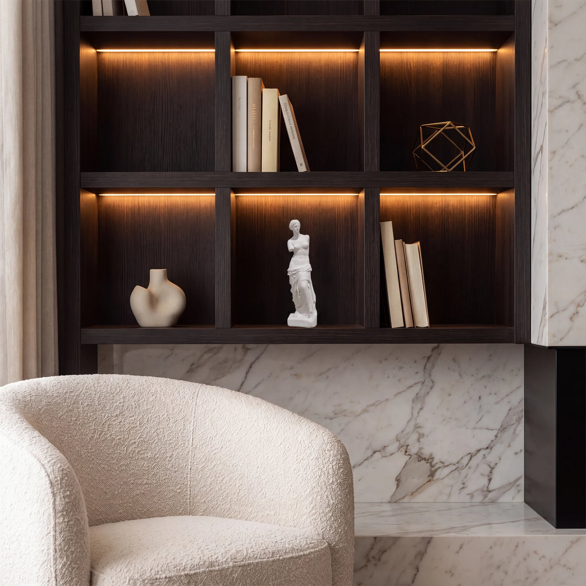 Escultura Vênus de Milo em gesso cru em estante escura de escritório com LED âmbar e poltrona bouclé