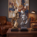 Escultura decorativa Silêncio e Paz dourada e prateada lado a lado em escritório com poltrona de couro e quadro abstrato