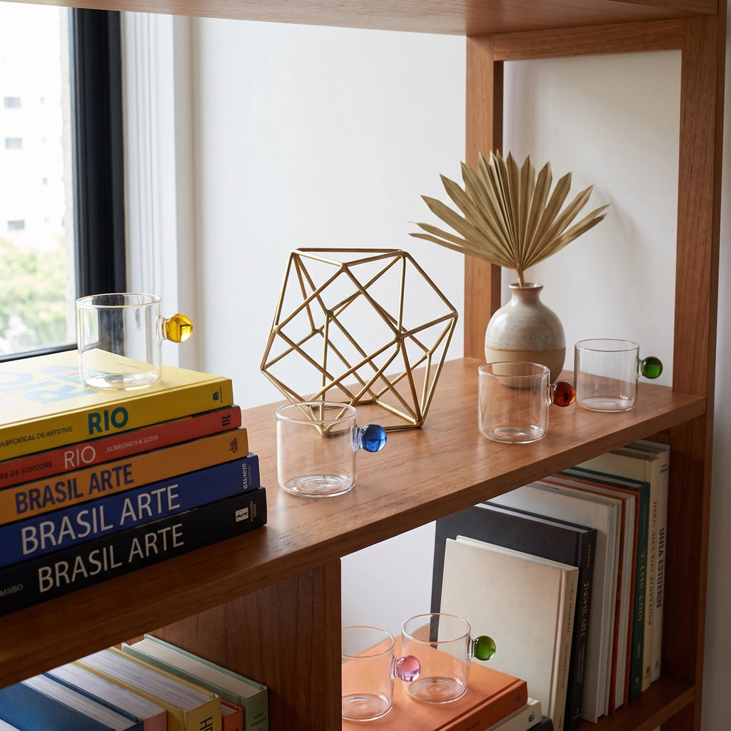 Xícaras Circo como decoração em estante com livros coloridos, objeto geométrico brass e palmeira seca