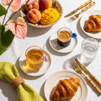 Brunch top-down com xícaras Circo, croissants, frutas tropicais e talheres dourados